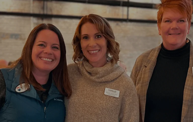 Two women pose for a photo in a festive, industrial-style venue, wearing name tags and cozy attire, with people mingling in the background.
