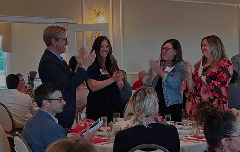A lively gathering in an elegantly decorated room with attendees clapping and celebrating, seated at tables adorned with dishes and drinks.