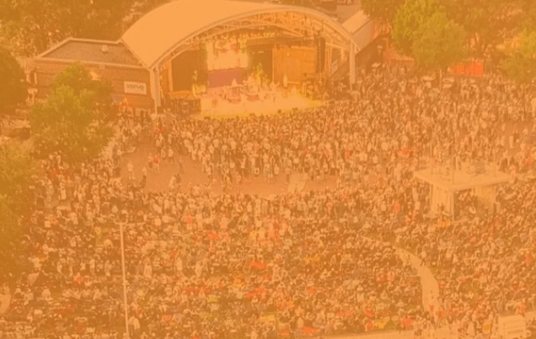 Aerial view of a large crowd gathered at an outdoor concert venue, with a stage and trees surrounding the audience.