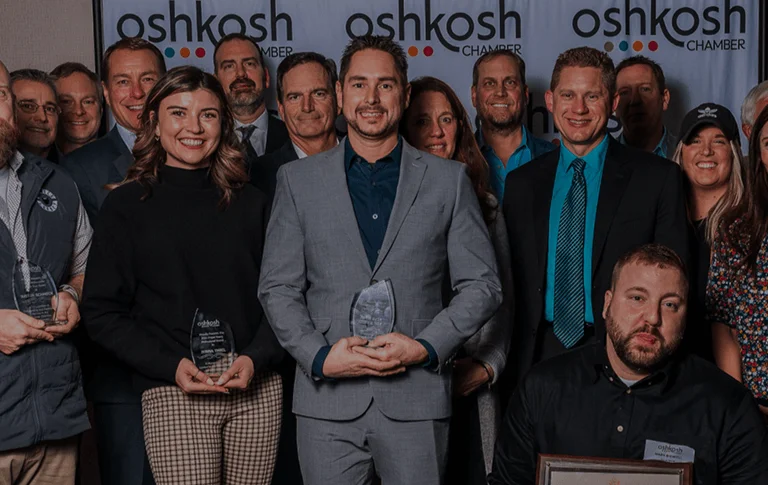 A group of award recipients poses together, holding trophies, in front of an Oshkosh Chamber banner at an awards ceremony.