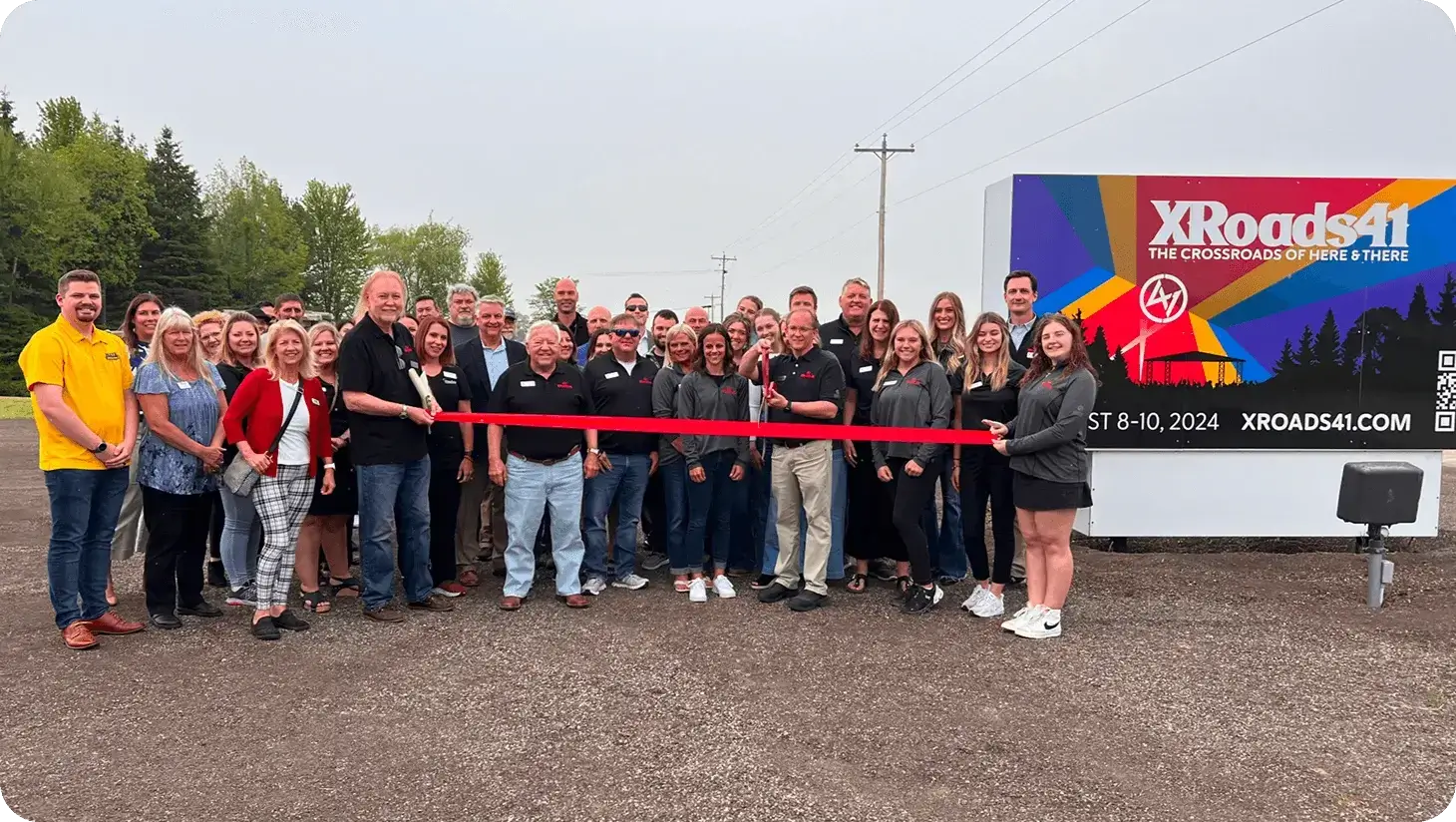 A group of people gathered for a ribbon-cutting ceremony in front of a colorful sign promoting XRoads41 event from August 8-10, 2024.