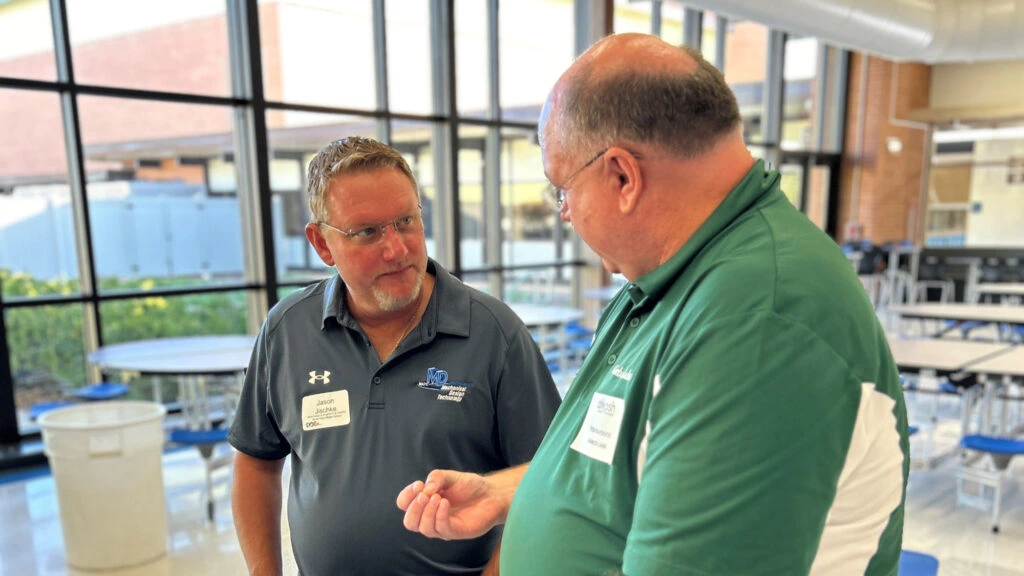 Two men wearing green shirts talking to each other in a bustling cafeteria