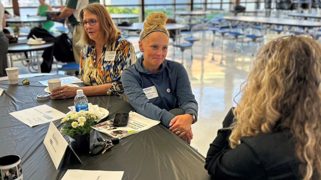 A group of three women sitting together at a table in a bustling cafeteria, sharing a meal and laughter